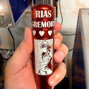 May include: A red metal container with a white graphic of a female anime character with the text "RIAS GREMORY" and "リアス" on the front.