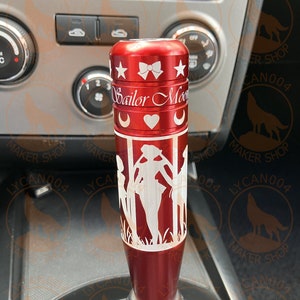 May include: Red metal gear shift knob with a white design of the Sailor Moon characters and the text "Sailor Moon".