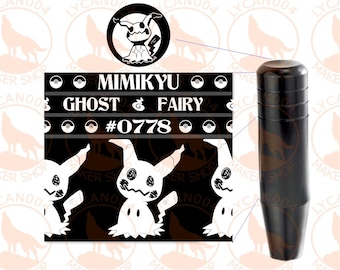 May include: A black cylindrical gear shift knob featuring a graphic print of the character Mimikyu. The design includes the text Mimikyu, Ghost, Fairy, and #0778, along with small circular icons resembling Poké Balls on a black background.