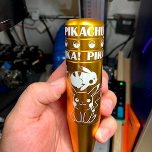 May include: A gold-colored cylindrical object with white text and graphics. The text reads "PIKACHU" and "KA! PIK". White illustrations of Pikachu and other Pokemon characters are visible. The object is held in a person's hand.
