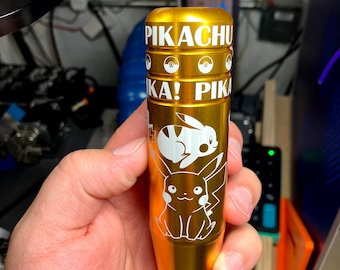 May include: A gold-colored cylindrical object with white text and graphics. The text reads "PIKACHU" and "KA! PIKA". White illustrations of Pikachu and a rabbit-like character are also visible. The object is held in a hand.