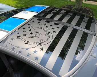 May include: A car roof featuring a graphic of an American flag with a stylized eagle head integrated into the star field. The design uses shades of black, grey, and white, with a glossy finish that reflects the surrounding environment and sky.