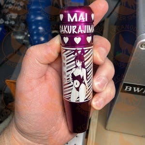 May include: A purple metal object with the name "MAI SAKURAJIMA" in white lettering and heart symbols. The object features a black and white illustration of a character in a bunny suit. The text "桜島麻衣" is also visible.