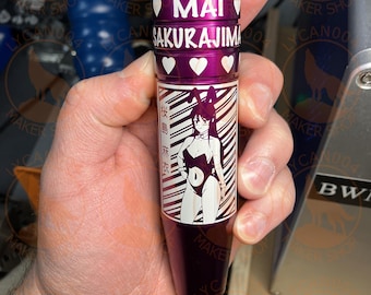 May include: A purple metal object with the name "MAI SAKURAJIMA" in white lettering and heart symbols. The object features a black and white illustration of a character in a bunny suit. The text "桜島麻衣" is also visible.