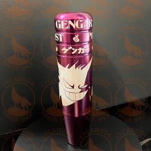 May include: A cylindrical, purple shift knob with white text that reads "GENGAR", "ST", and "PO". The knob features a white graphic of a smiling ghost-like character and Japanese text. The background is a dark surface with a repeating logo.