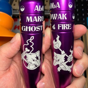 May include: Two purple metal objects with white text and illustrations. The text includes "ALOLA", "MAROWAK", "GHOST", "OWAK", and "FIRE". Each object features a white illustration of a cartoon character. The objects are held in a person's hand.