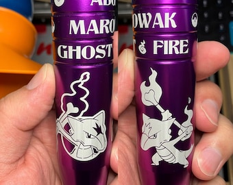 May include: Two purple metal objects with white text and illustrations. The text includes "ALOLA", "MAROWAK", "GHOST", "OWAK", and "FIRE". Each object features a white illustration of a cartoon character. The objects are held in a person's hand.