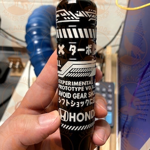 May include: Black and white Honda shift knob with Japanese text and the words "EXPERIMENTAL PROTOTYPE VO.1" and "AVOID GEAR SHOCK". The shift knob has a cylindrical shape and is held in a hand.