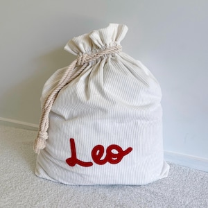 May include: A white corduroy drawstring sack featuring the name Leo in red script lettering. The bag is cinched at the top with a thick, twisted beige rope cord that is knotted on the side, creating a gathered ruffle effect at the opening.