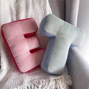 May include: Two corduroy letter-shaped cushions in the form of the letters E and T. The letter E is light pink with a dark red border, while the letter T is light green with a light blue border. Both cushions feature a ribbed fabric texture.