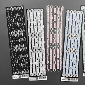 May include: Five sheets of sticker paper with different designs. Each sheet has a repeating pattern of a heart-shaped buckle with a chain link and a row of small circles. The sheets are white, pink, light blue, and green.