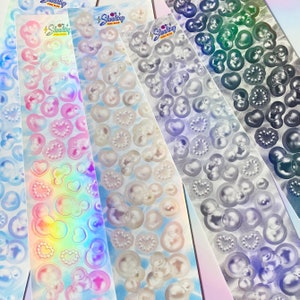 May include: Four rolls of iridescent holographic stickers featuring various pearl-like shapes, including hearts, circles, and Mickey Mouse ears. The stickers are on a white background with a light blue cloud pattern. The text "Shocking Pink Rose" is printed on each roll.