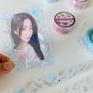K&ouml;nnte beinhalten: Ein transparenter Plastik-Bilderrahmen mit holografischem Design mit blauen Herzen, Wolken und Sternen. Der Rahmen ist mit einem glitzernden Finish verziert. Der Text "I SHOCKING PINK ROSE" ist auf einer rosa-wei&szlig;en Rolle Washi-Tape sichtbar.