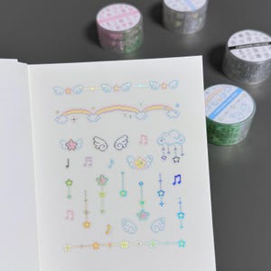 May include: A notebook page with pixelated stickers featuring rainbows, clouds, stars, musical notes, and angel wings. Several rolls of decorative tape with various designs are in the background.