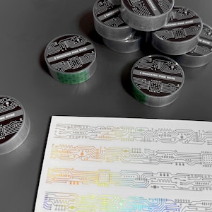 Peut inclure: Plusieurs rouleaux de ruban adhésif décoratif argenté avec un motif de circuit imprimé et le texte "SHOCKING PINK ROSE". Le cœur du ruban est vert. Une feuille d'autocollants avec un motif similaire est également présente.