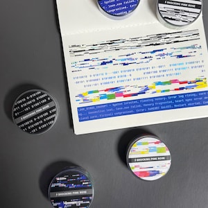 Peut inclure: Plusieurs autocollants ronds avec un motif de glitch numérique et le texte "SHOCKING PINK ROSE." Les autocollants ont un schéma de couleurs noir, blanc et rose. Un morceau de papier avec du code binaire et des messages d'erreur est également visible.