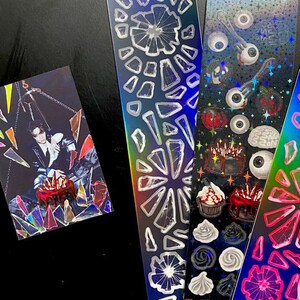 May include: Three holographic sticker sheets with various designs. The first sheet features a pattern of broken glass shards. The second sheet features a pattern of eyes, brains, and cakes. The third sheet features a pattern of colorful swirls.