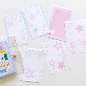 May include: A collection of pastel-colored stationery items, including notepads and a clear binder. The notepads feature a star pattern in pink and blue, with some having lined pages. The binder has decorative stickers.