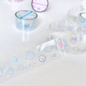 K&ouml;nnte beinhalten: Eine Rolle transparentes, schimmerndes Washi-Tape mit einem sich wiederholenden Muster aus blauen und rosa pixelartigen Blasen. Das Band liegt auf einer wei&szlig;en Oberfl&auml;che.