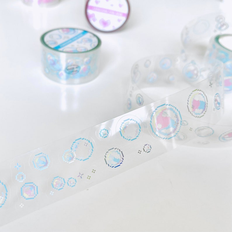 K&ouml;nnte beinhalten: Eine Rolle transparentes, schimmerndes Washi-Tape mit einem sich wiederholenden Muster aus blauen und rosa pixelartigen Blasen. Das Band liegt auf einer wei&szlig;en Oberfl&auml;che.
