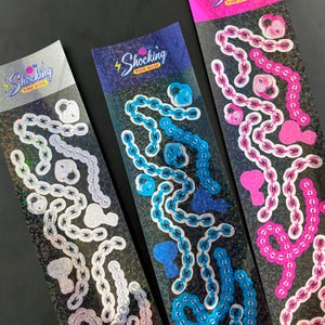 Peut inclure: Trois planches d'autocollants avec des motifs de chaînes et de cadenas argentés, bleus et roses. Chaque planche porte le texte "Shocking Pink Rose". Les autocollants ont un effet holographique sur fond noir.