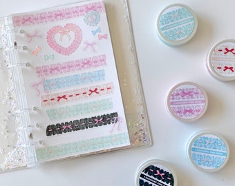 May include: A clear plastic binder with a white sheet of paper inside. The paper has several rows of pink, blue, and black lace trim stickers. There are also several small stickers of hearts and bows. The binder is on a white surface with a scattering of glitter. There are six small round containers with lids on the surface. The lids are decorated with pink, blue, and black lace trim and the text "Shocking Pink Rose".