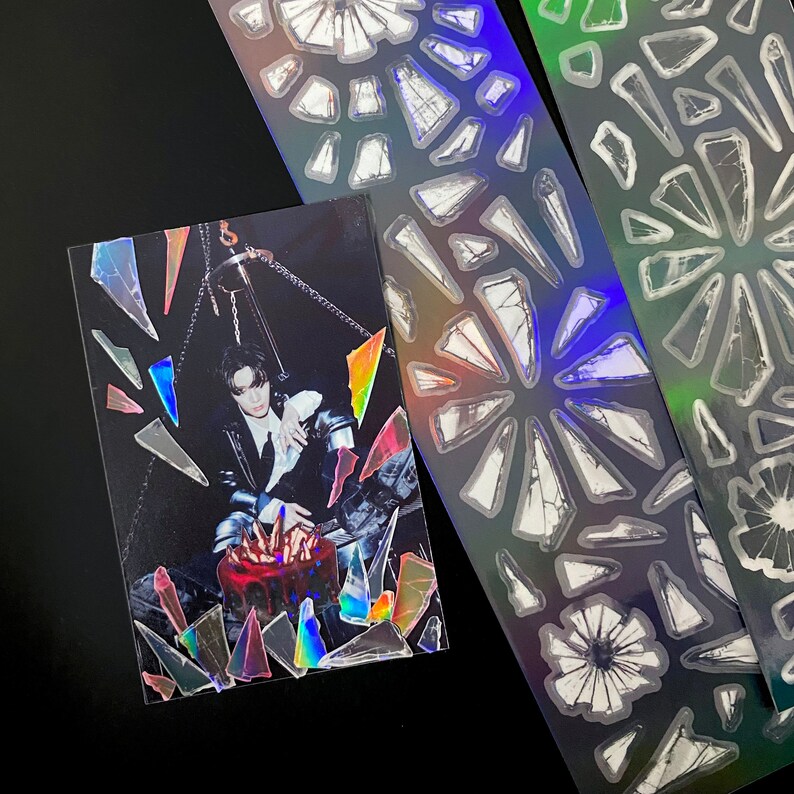 May include: A black and white photo of a person wearing a black jacket and holding a red and white cake. The photo is surrounded by iridescent holographic stickers in the shape of broken glass.