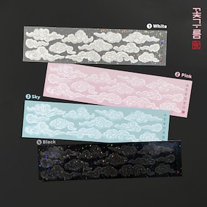 May include: Four sheets of cloud stickers with different coloured backgrounds. The first sheet is white with iridescent glitter, the second sheet is pink, the third sheet is light blue, and the fourth sheet is black with iridescent glitter. Each sheet has a variety of cloud shapes and sizes.