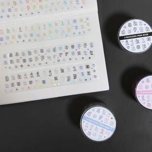 Peut inclure: Présentation d'autocollants alphabet et de rouleaux de ruban décoratifs. Les autocollants, avec des lettres minuscules et des accents holographiques, sont sur des bandes transparentes. Les rouleaux de ruban portent l'inscription "SHOCKING PINK ROSE" avec l'alphabet.