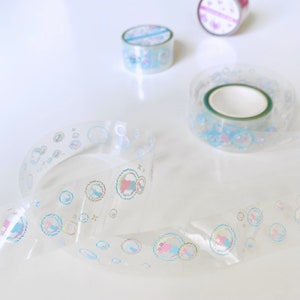 K&ouml;nnte beinhalten: Drei Rollen transparentes Washi-Tape mit schimmernden Blasenmustern. Das Klebeband liegt auf einer wei&szlig;en Oberfl&auml;che.