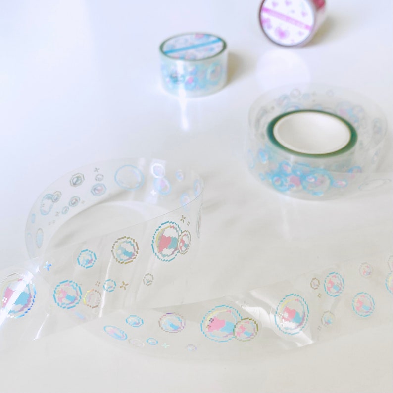 K&ouml;nnte beinhalten: Drei Rollen transparentes Washi-Tape mit schimmernden Blasenmustern. Das Klebeband liegt auf einer wei&szlig;en Oberfl&auml;che.