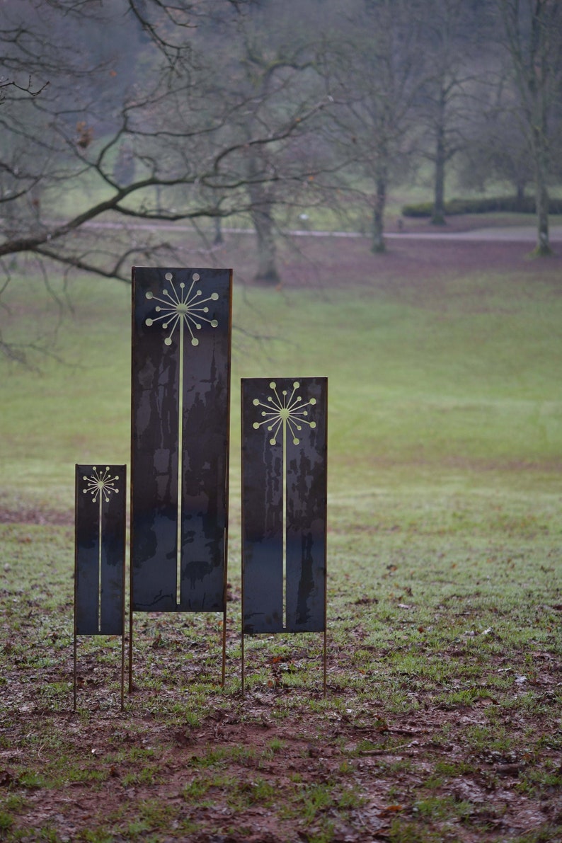 May include: Three metal garden sculptures in the shape of stylized flowers. The sculptures are different sizes and are arranged in a row on a grassy lawn.