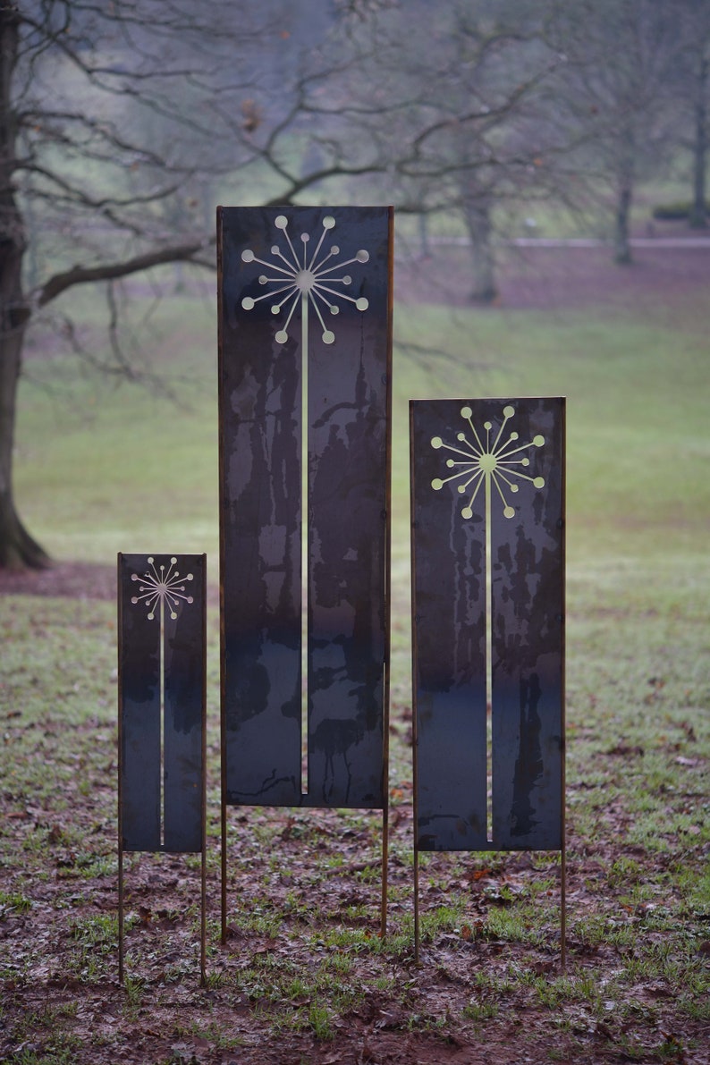 May include: Three metal garden stakes with cut-out dandelion designs. The stakes are different heights and are placed in a grassy area.