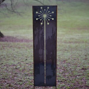 May include: A tall, metal garden sculpture with a cut-out design of a stylized flower. The sculpture is painted a dark gray color and has a rusty patina. It is standing on a bed of brown dirt and green grass.