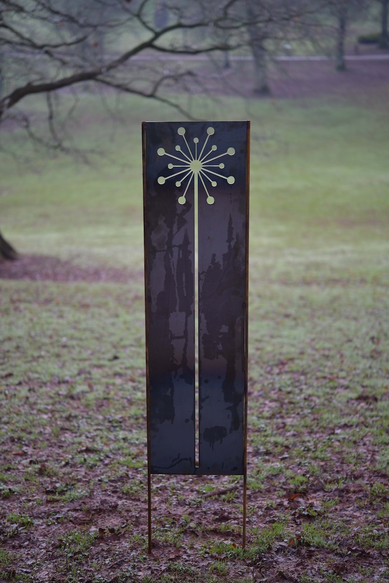 May include: A tall, metal garden sculpture with a cut-out design of a stylized flower. The sculpture is painted a dark gray color and has a rusty patina. It is standing on a bed of brown dirt and green grass.