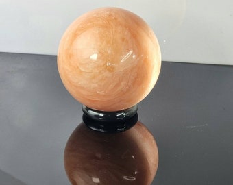 Könnte beinhalten: Eine große, glatte, orangefarbene und weiße Kugel aus poliertem Stein. Die Kugel steht auf einem schwarzen Sockel.