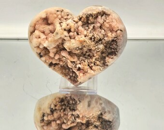 May include: A heart-shaped pink and brown geode with a rough, textured surface. The geode is displayed on a clear stand.