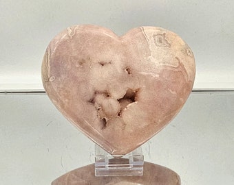 May include: A heart-shaped pink and white agate geode with a crystal formation inside. The geode is on a clear stand.