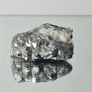 Può includere: Un campione minerale ruvido, grigio e argentato, con una superficie riflettente. Il minerale ha una struttura cristallina ed è una formazione rocciosa naturale.