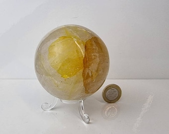 Puede incluir: Una esfera de cuarzo transparente con inclusiones amarillas, expuesta en un soporte transparente. La esfera es aproximadamente del tamaño de una moneda de 1 euro.