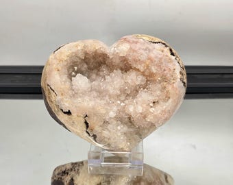 May include: A heart-shaped geode with a crystalline interior. The exterior is a mix of beige and brown, with dark veins. The inside is filled with sparkling, light-colored crystals. The geode is displayed on a clear stand.
