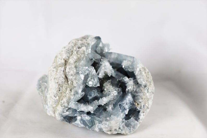 May include: A cluster of blue celestite crystals in a natural rock formation. The crystals are translucent and have a light blue color. The rock is gray and has a rough texture.
