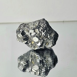 Può includere: Una roccia grigia ruvida con un gruppo di cristalli argentati lucenti incastonati. I cristalli hanno una forma geometrica sfaccettata.