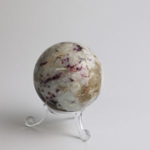 May include: A white and grey sphere with red and black markings, displayed on a clear plastic stand.