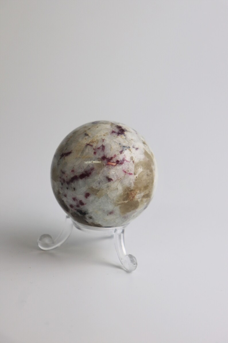 May include: A white and gray sphere with red and black markings, displayed on a clear plastic stand.