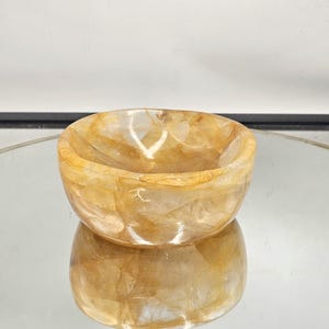 May include: A small, round bowl made of clear, yellow-tinted quartz crystal. The bowl has a smooth, polished surface and is perfect for holding small items.