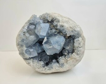 May include: A geode with a large opening revealing a cluster of light blue crystals inside. The geode is a light gray color and has a rough, textured surface.
