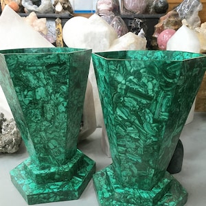May include: Two green malachite vases with a geometric design. The vases have a hexagonal base and a flared, faceted body. The malachite has a swirling pattern of dark and light green. The vases are displayed on a light gray surface.