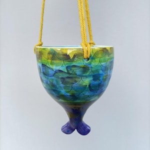 May include: A hanging ceramic planter with a mermaid tail design. The planter is painted with a watercolor effect in shades of blue, green, and yellow. It is suspended by yellow rope.