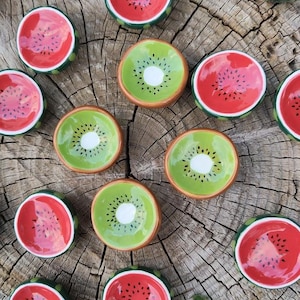 Op de afbeelding: Een verzameling kleine keramische kommen die ontworpen zijn om op plakjes watermeloen en kiwi te lijken. De watermeloenkommen hebben rode binnenkanten met zwarte zaaddetails en groene randen. De kiwikommen hebben groene binnenkanten met een wit centrum en bruine randen. De kommen zijn gerangschikt op een houten oppervlak.